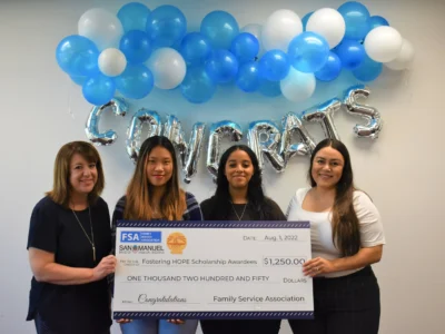 Four women smile holding a $1,250 scholarship check, with "CONGRATS" balloons and blue balloons behind them.