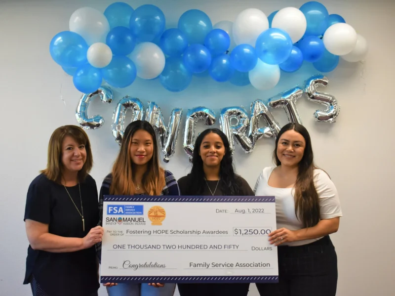 Four women smile holding a $1,250 scholarship check, with "CONGRATS" balloons and blue balloons behind them.