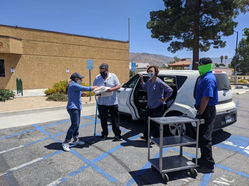 Four people wearing masks stand by a car in a parking lot, handling papers next to a metal cart.
