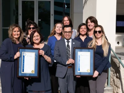 A group of people pose outside, two in front holding framed certificates and smiling at the camera.