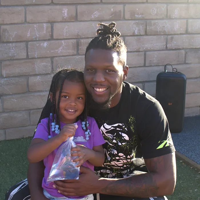 Smiling man and young girl with braids pose together outside; the girl holds a clear bag of snacks.