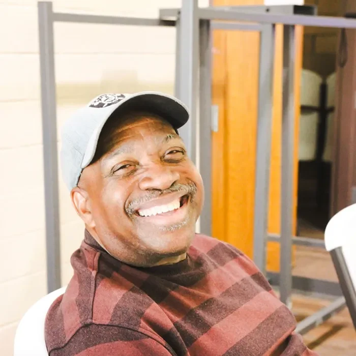 Smiling man in a cap and striped shirt sitting indoors near a table and chairs.