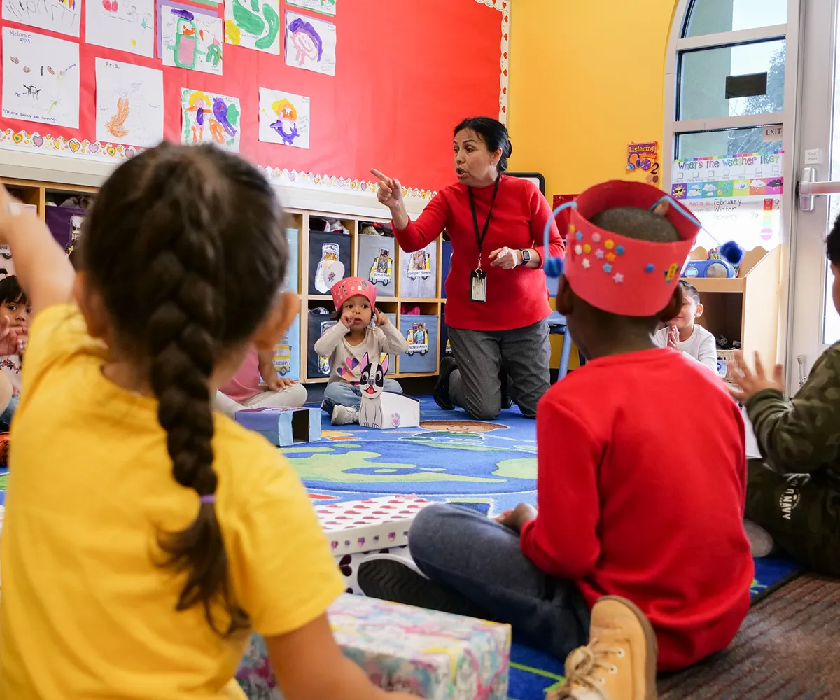 A teacher talks to young children sitting on the floor in a colorful classroom, some raising their hands.