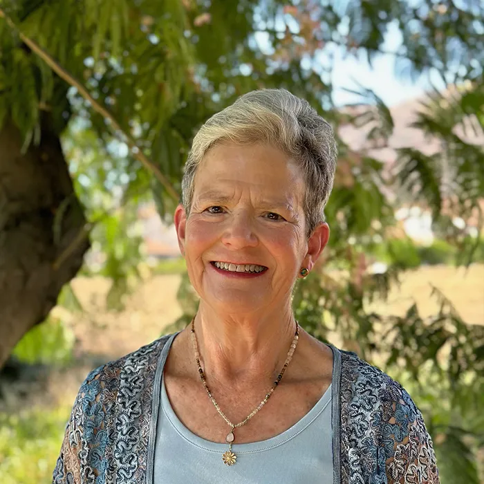 Smiling older woman with short gray hair stands outdoors in front of green trees, wearing a blue top.