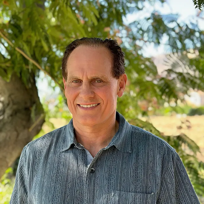 Smiling man with short brown hair stands outdoors in front of green trees, wearing a patterned gray shirt.