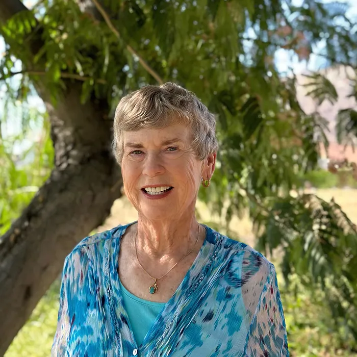 Smiling older woman with short gray hair stands outdoors by a tree, wearing a blue patterned top.