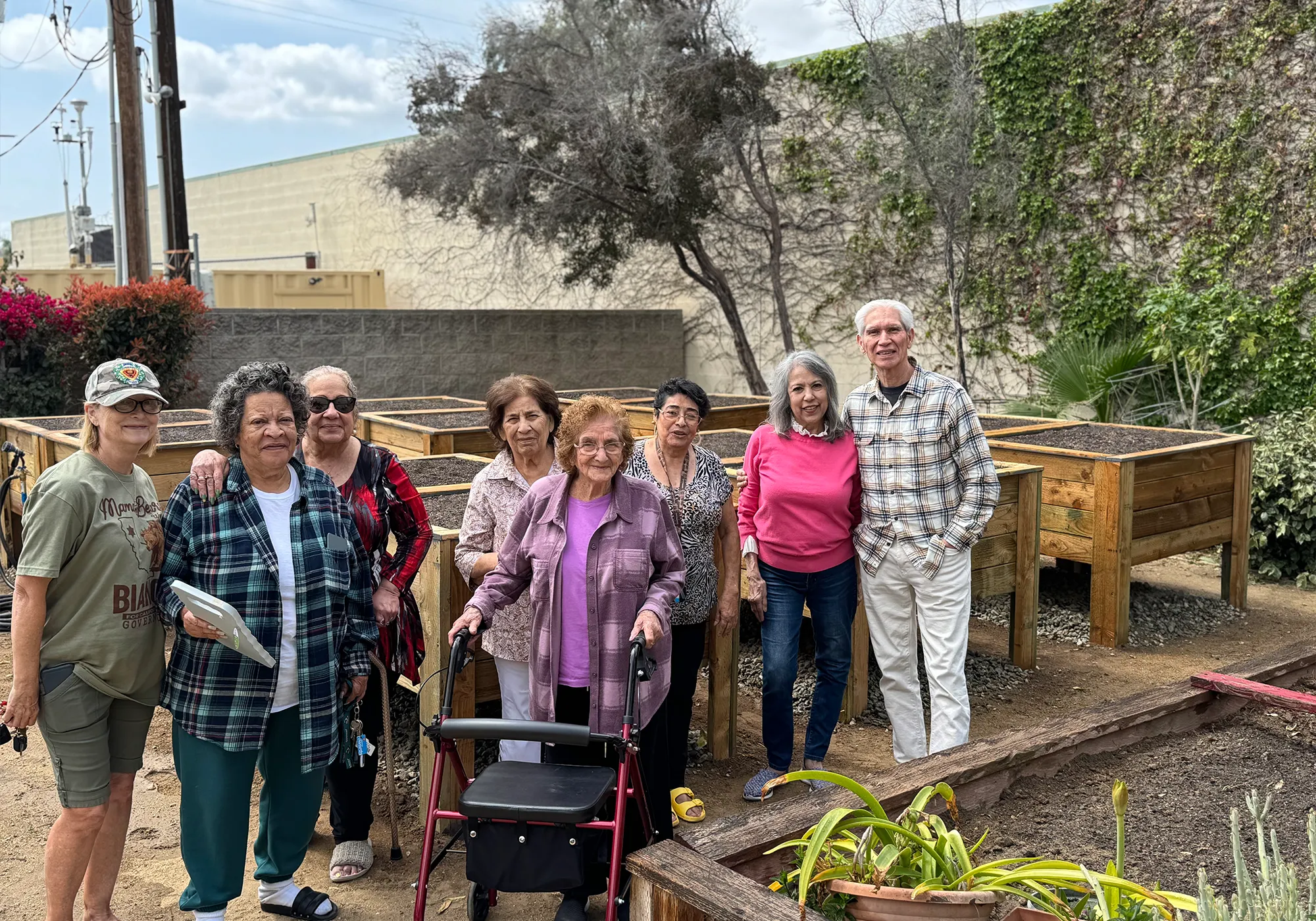 A group of smiling seniors poses together in a community garden with raised planting beds.