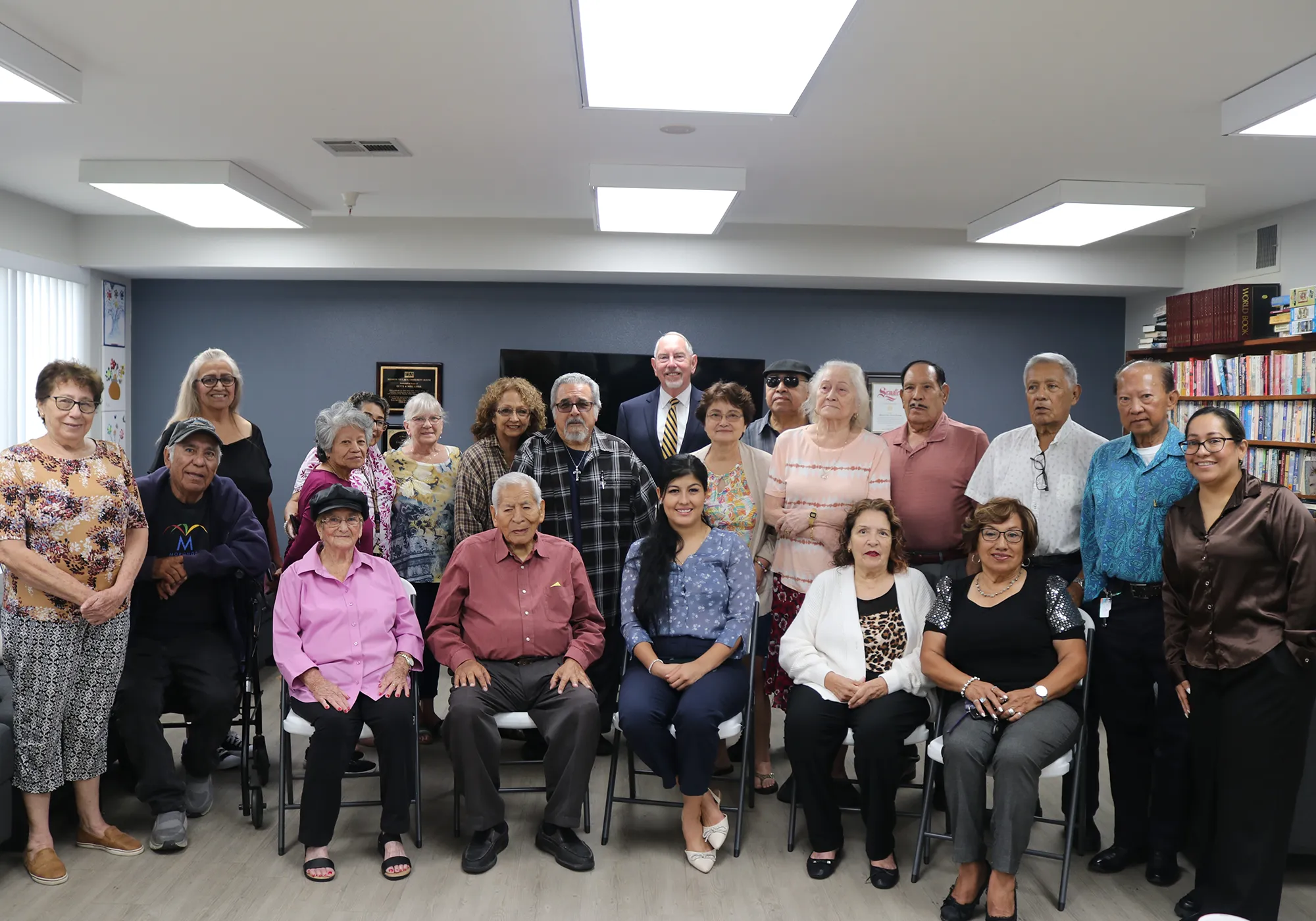 A group of older adults and a few younger people pose together and smile in a library or community room.