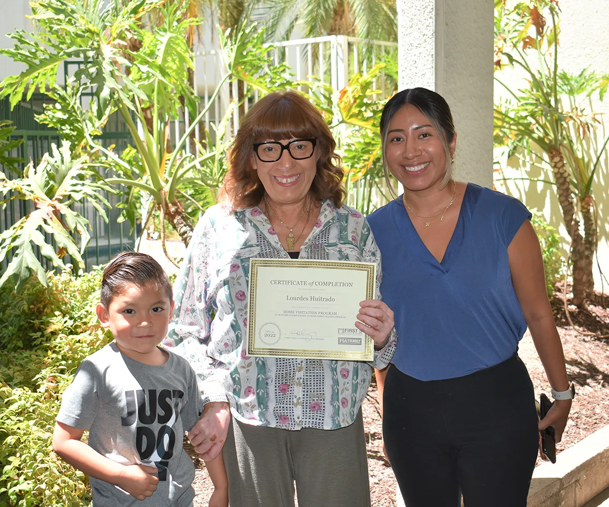 Three people smile outdoors; the middle person holds a certificate of completion.