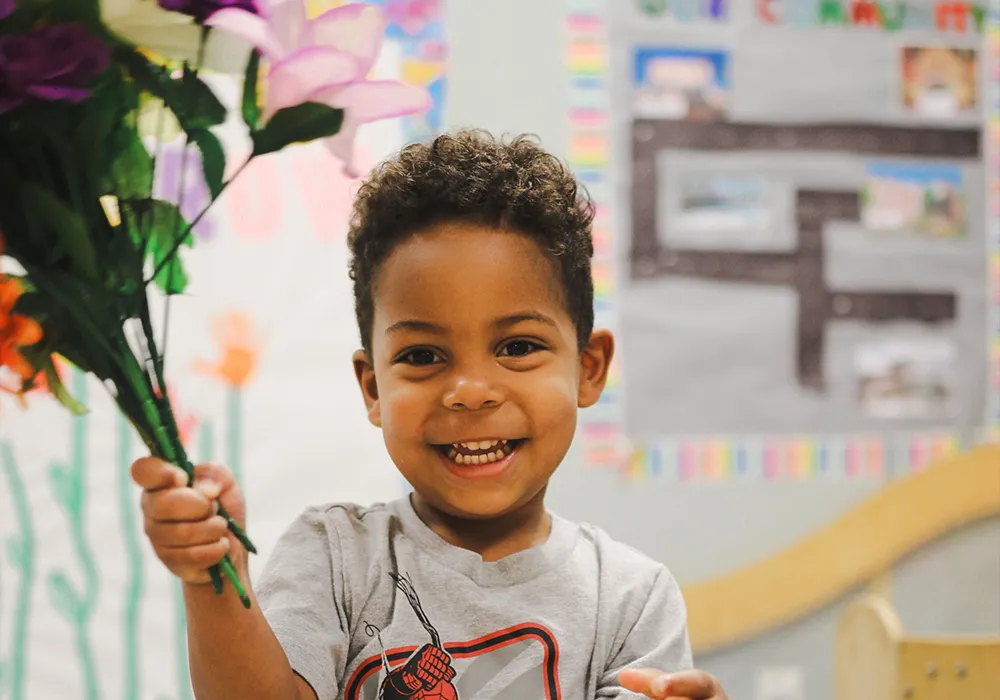 Smiling young boy holds a bouquet of colorful flowers in a bright classroom setting.