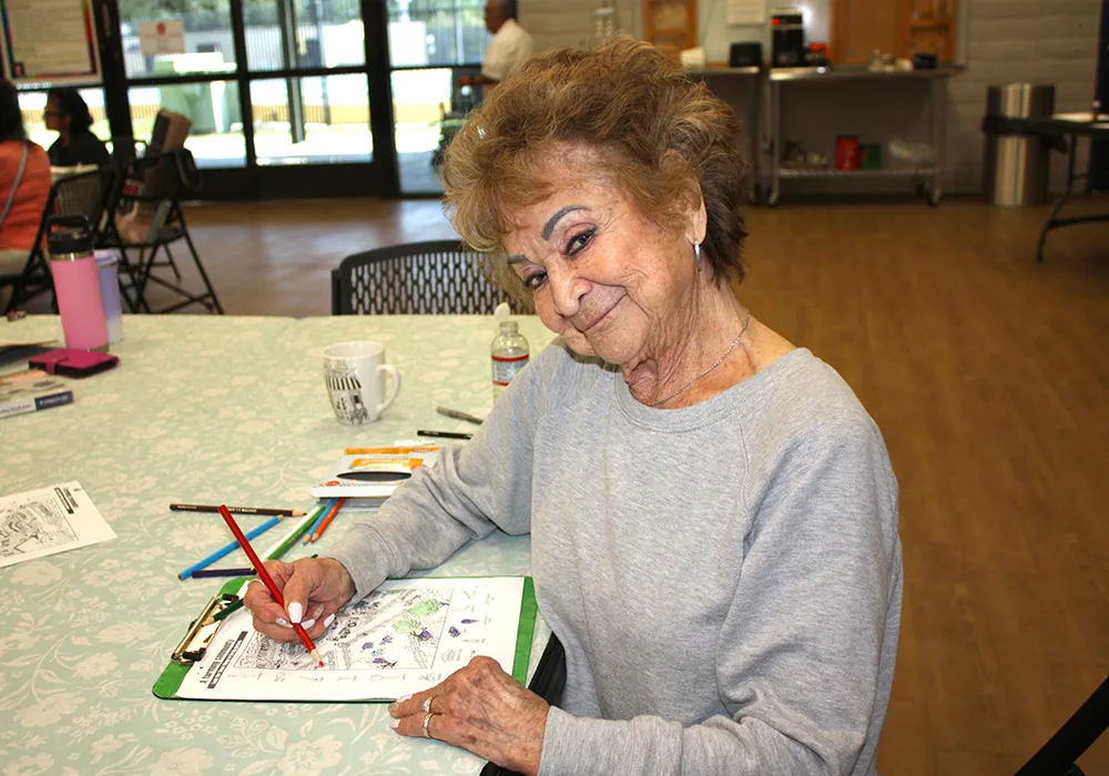 Smiling elderly woman sitting at a table, coloring a worksheet with colored pencils in a community room.