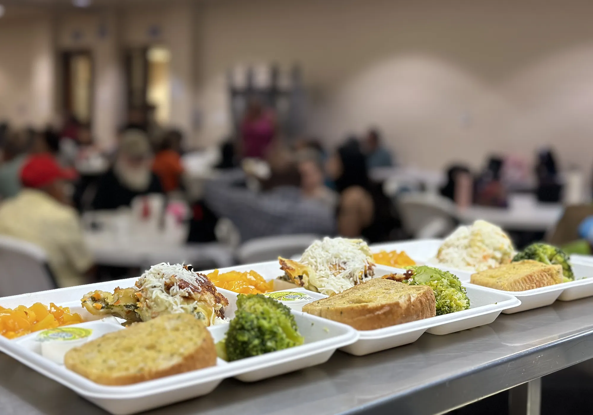 Trays of food with bread, broccoli, pasta, and fruit are lined up, with people seated in the background.