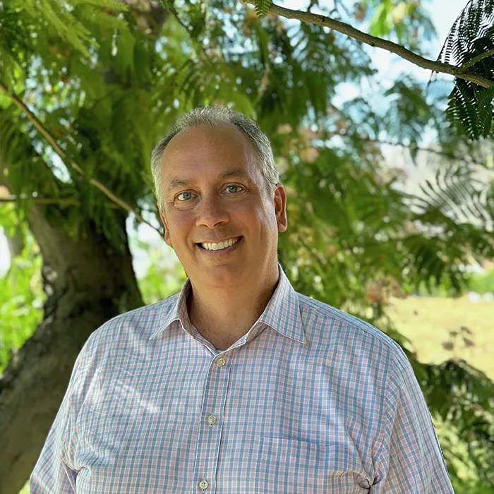 Smiling man in a pink checked shirt standing outdoors in front of green trees and sunlight.