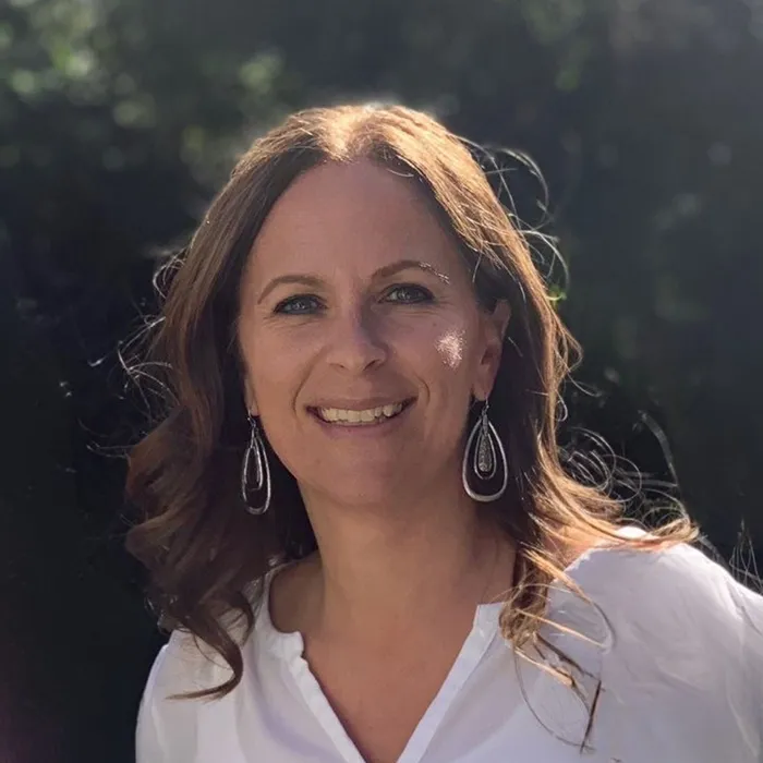 Woman with wavy brown hair and white blouse smiling outdoors, sunlight in the background.