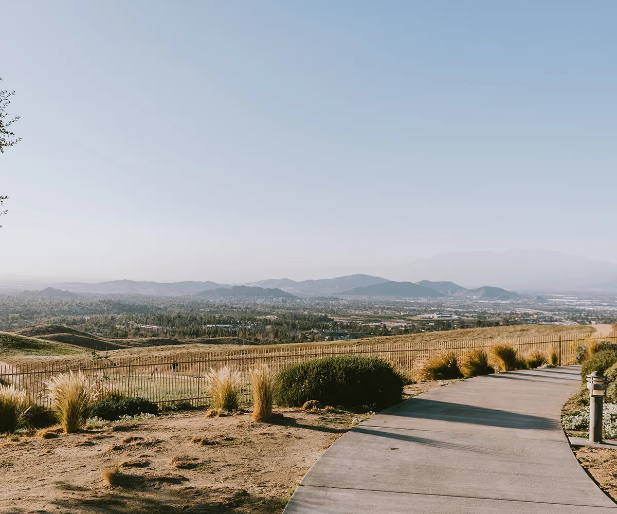 Curved path beside a fence overlooks grassy hills, cityscape, and distant mountains under a clear sky.