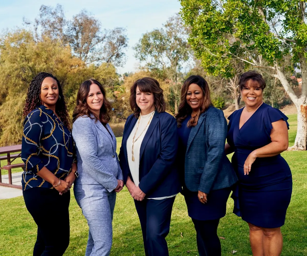 Five women in business attire standing together and smiling outdoors in a grassy park setting.