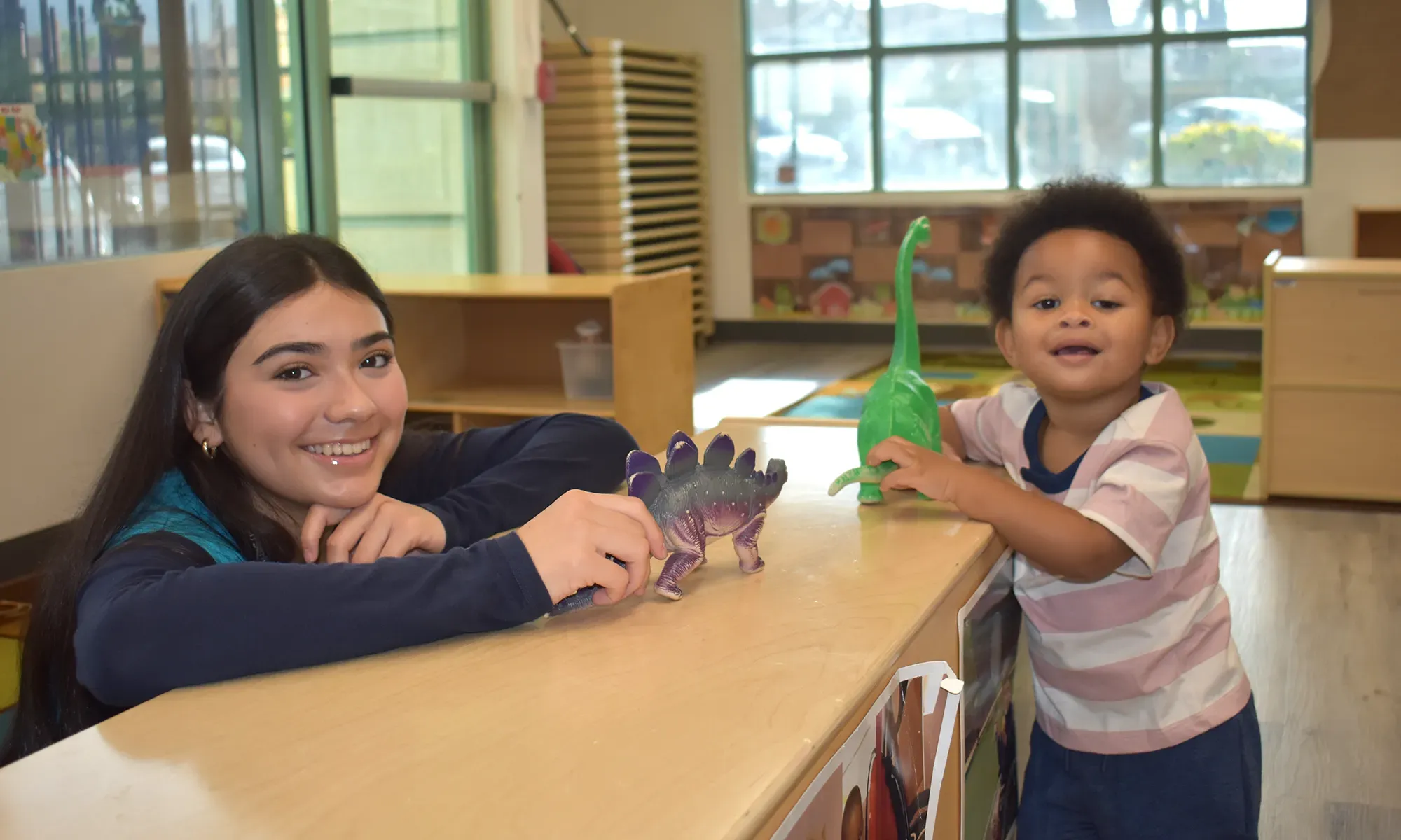 A woman and a young boy smile while playing with dinosaur toys at a table in a bright classroom.