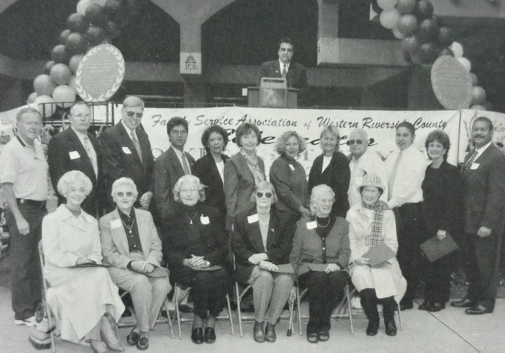 A group of adults pose in front of a banner and balloon arch at an outdoor event; many are wearing name tags.