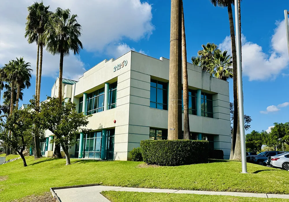 Two-story modern office building with large windows, palm trees, and green grass under a blue sky.