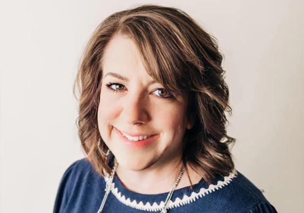 Smiling woman with wavy brown hair wearing a navy blue top and necklace, posing against a light background.
