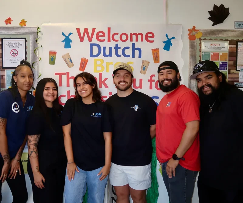Six smiling people standing together in front of a colorful "Welcome Dutch Bros" sign on a classroom wall.