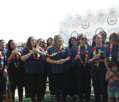A group of women in navy shirts and colorful leis stand together, smiling at an outdoor event.
