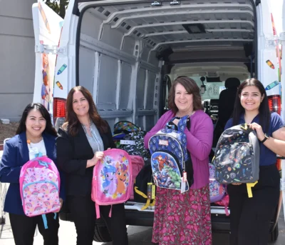 Four women stand by an open van holding colorful backpacks filled with school supplies.