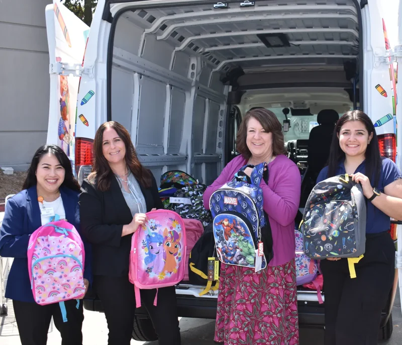 Four women stand by an open van holding colorful backpacks filled with school supplies.